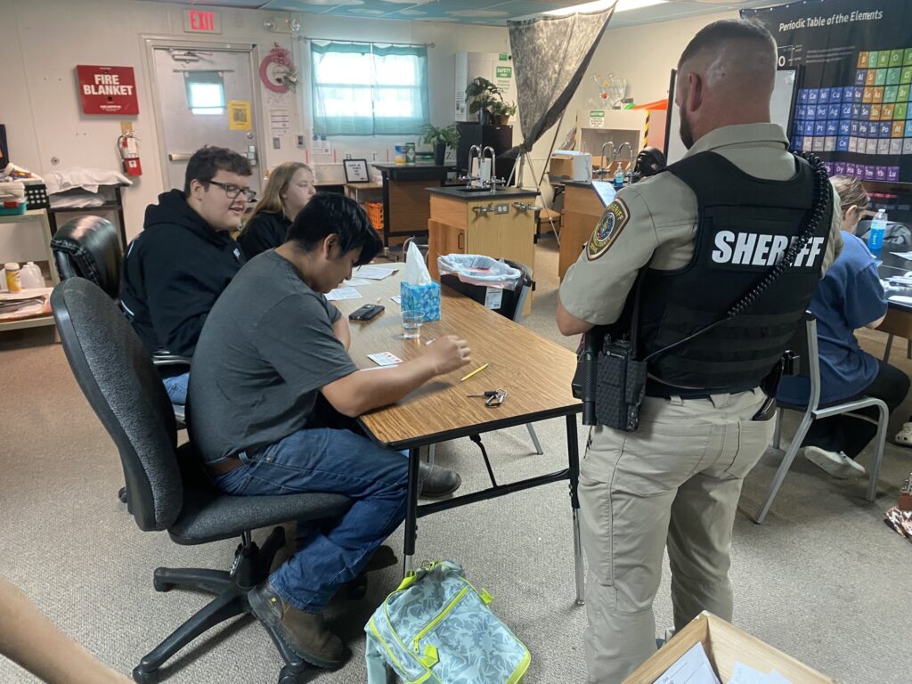 Students sit at a table while the campus patrol officer presents information. The officers back is to the camera.