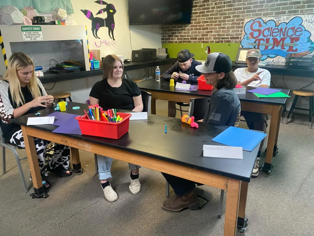 a group of students take part in first date craft activities at Brazos River Charter School. In the foreground, three students said that the table with a bright red container full of various colored pens. In the background, one student is completing a project while the second student looks at the camera. Behind that student is a homemade sign with a blue cloud that reads Science Time in red lettering.