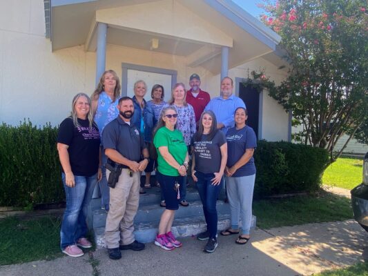 The staff of Brazos River Charter School standing in three rows. The first row shows for teachers and the on-site police officer. The second row shows for teachers. The third row shows two teachers.
