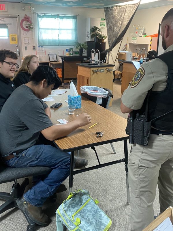 Students sit at a table while the campus patrol officer presents information. The officers back is to the camera.